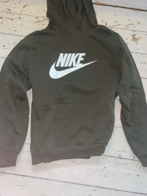 Mörkgrön Nike hoodie - Snygg mörkgrön hoodie från Nike med stor vit logga på bröstet. Hoodien har huva och ribbade muddar vid ärmslut och nederkant. Tillverkad i mjukt material som känns skönt mot huden. Perfekt för en avslappnad och sportig stil.
