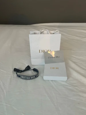 Blått Christian Dior armband - Säljer ett snyggt blått armband från Christian Dior med broderad logga och justerbar knytning. Armbandet har dekorativa tofsar i ändarna och levereras med originalpåse och ask. Perfekt accessoar för att lyfta din outfit.