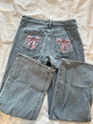 Ljusblå jeans med rosa rosetter - Snygga ljusblå jeans med raka ben och två bakfickor dekorerade med broderade rosa rosetter. Jeansen har klassisk femficksdesign och stängs med knapp och dragkedja. Perfekta för dig som vill ha en söt och trendig detalj på dina jeans.
