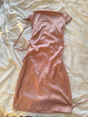 Rosa satinklänning med snörning - Säljer en snygg rosa satinklänning i midilängd med axelbandslös design och trendig snörning i ryggen. Klänningen har en slät, glansig yta och figurnära passform. Perfekt för dig som vill ha en chic och modern look.