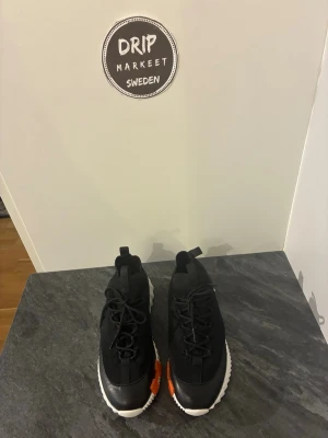 Hermees sneakers svarta  - Snygga svarta sneakers med stickad ovandel, vita sulor och en cool orange detalj på sidan. Skorna har rund tå, svarta skosnören och dragflikar både fram och bak för enkel påtagning. Perfekta för dig som vill ha en modern och sportig look.
