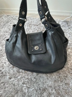 Marc by Marc Jacobs väska - Säljer nu min fina Marc by Marc Jacobs-väska i svart läder och silverdetaljer i mycket fint och välbevarat skick. En klassisk och rymlig modell som passar både till vardag och arbete. Jag har anvant den som skolväska, och den rymmer utan problem A4-block och en laptop. Säljs med tillhörande dammpåse. Ett av magnetfästena på en av sidfickorna har något svagare magnetfunktion, men i övrigt är allt som det ska.