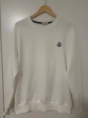 Vit sweatshirt från Moncler - Klassisk vit sweatshirt från Moncler med rund halsringning och diskret Moncler-logga broderad på bröstet. Tröjan har ribbade muddar vid ärmslut och nederkant, samt en avslappnad passform. Perfekt för en clean och stilren look.