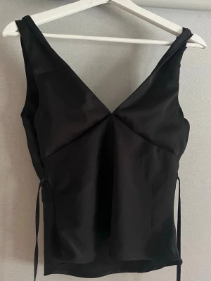 Svart omlottopp med v-ringning - Snygg svart omlottopp med djup v-ringning och knytband i sidan. Toppen är ärmlös och har en elegant, figurnära passform. Perfekt att styla med smycken för en trendig look.