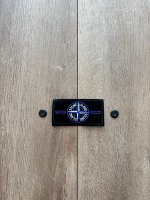 Stone Island Patch / Badge – Svart med blå/lila brodyr & knappar - Säljer en klassisk Stone Island-badge i svart med blå/lila brodyr, inklusive tillhörande originalknappar precis som på bilderna. Patchen är helt ny, oanvänd och i perfekt skick. Passar utmärkt som reservdel eller tillbehör till din Stone Island-jacka eller tröja. 