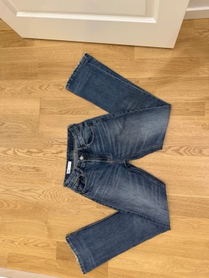 Blå flare jeans från ZARA - Säljer ett par klassiska blå jeans från ZARA med flare passform och fem fickor. Jeansen har en snygg tvättad look med subtila slitningar och kontrastsömmar. Tillverkade i bomull för en skön känsla och tidlös stil.