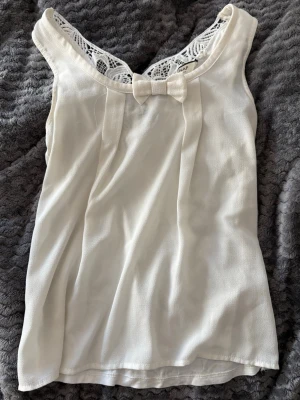 Söt vit ärmlös topp med spetsdetaljer - Söt vit ärmlös topp med spets i halsringningen och en liten rosett framtill. Toppen har ett luftigt tyg och vackra spetsmönster både fram och bak, vilket ger en romantisk vibe. Perfekt att matcha med jeans eller kjol för en fräsch look.