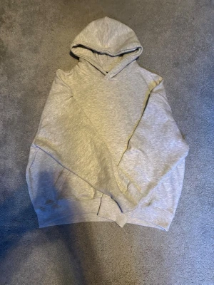 Ljusgrå basic hoodie med huva från pull and bear - En enkel och stilren ljusgrå hoodie med huva och magficka. Hoodien har ribbade muddar vid ärmslut och nederkant, samt en klassisk passform som funkar till det mesta. Perfekt för dig som gillar en clean och avslappnad look.