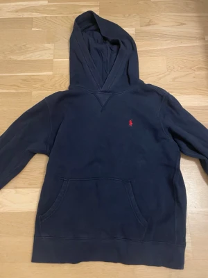 Mörkblå hoodie med röd logga - Snygg mörkblå hoodie med luva och ficka framtill. På bröstet finns en liten röd broderad logga. Hoodien har långa ärmar och ribbade muddar vid ärmslut och nederkant. Perfekt för en avslappnad och stilren look.