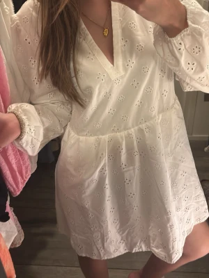 Vit kort klänning med broderade blommor - klänning med v-ringning och långa ärmar. Klänningen har broderade blommor över hela tyget och är gjord i lätt bomull. 