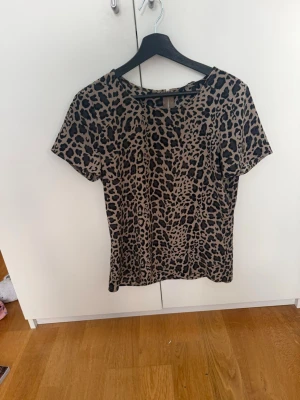 Leopardmönstrad t-shirt med dragkedja - Snygg t-shirt/blus med leopardmönster i beige och svart. Modellen har korta ärmar och en dragkedja i ryggen. 