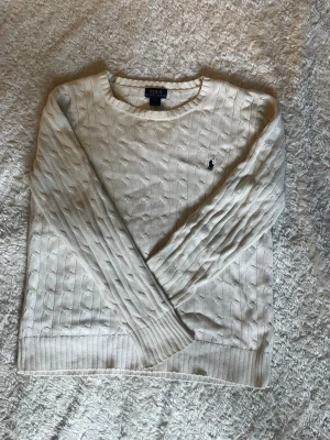 Vit kabelstickad tröja Polo Ralph Lauren - Kabelstickad vit tröja från Polo Ralph Lauren med rund halsringning och diskret broderad logga på bröstet. Tröjan har långa ärmar och ribbade muddar vid ärmslut och nederkant. Perfekt för en klassisk och stilren look.
