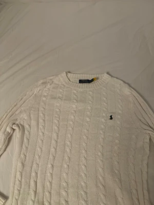Vit kabelstickad tröja Polo Ralph Lauren - Skulle passa dig som är 175-180 cm. Kabelstickad vit tröja från Polo Ralph Lauren med rund halsringning och den klassiska broderade loggan på bröstet. Tröjan har långa ärmar och ribbade muddar. Perfekt för dig som gillar en clean och tidlös stil.
