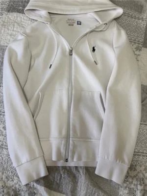 POLO RALPH LAUREN - Säljer en vit hoodie med dragkedja och huva. Hoodien har fickor framtill och en liten broderad logga på bröstet. Tillverkad i mjukt material och har dragsko i huvan. Perfekt för en avslappnad och stilren look.