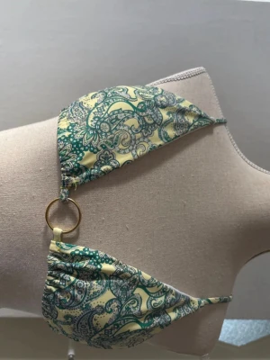 Gul och grön bikini med paisleymönster - Trendigt bikiniöverdel i gult och grönt med paisleyinspirerat mönster. Triangelformade kupor och smala axelband, dekorerad med en guldfärgad ring i mitten. Perfekt för sommarens strandhäng.
