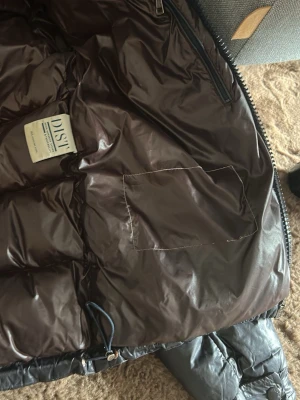 Svart dunjacka med dragkedja - Snygg svart dunjacka med glansig finish och quiltad design. Jackan har dragkedja framtill, innerficka och detaljer i mörkbrunt foder. Perfekt för kalla vinterdagar och håller dig varm med äkta dunfyllning. Jackan har några hål och sönder dragkedja men den går fortfarande att stänga