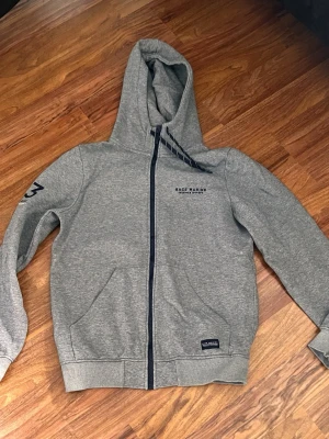 Grå zip-hoodie från Sail Racing - Snygg grå hoodie med dragkedja från Sail Racing. Har huva med snören, två fickor fram och tryck på bröstet samt ärmen. Tillverkad i mjukt sweatshirtmaterial, perfekt för chill dagar. Marinblå detaljer vid dragkedjan och logga på bröstet.