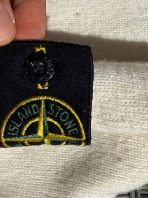 Vit stickad tröja från Stone Island - Vit stickad tröja från Stone Island i ull med klassisk svart och gul logotyp-patch på ärmen. Tröjan har rund halsringning och är ribbstickad vid hals, ärmslut och nederkant. Perfekt för dig som gillar stilrena och exklusiva plagg. Tröjan är i använd skick så de bilderna!