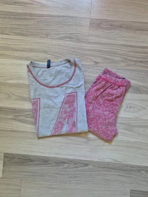 Grå och rosa pyjamas med siffror - Mysig pyjamas med grå t-shirt och rosa detaljer. T-shirten har siffran 14 i rosa spetsmönster framtill och matchande sömmar. Byxorna är rosa med vitt spetsinspirerat mönster och resår i midjan. Perfekt för slappa dagar hemma.