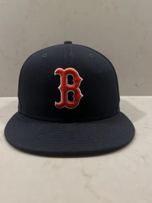 Mörkblå Boston Red Sox keps - Snygg mörkblå keps med röd och vit Boston Red Sox-logga framtill. Klassisk flat brim-modell och broderad detalj på framsidan. Perfekt för dig som gillar sportig stil och vill representera ditt favoritlag. Pris kan diskuteras 