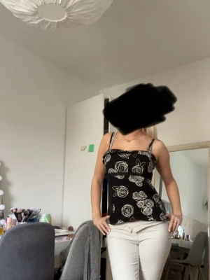 Svart blommig linnetopp med smala band - Trendig svart linnetopp med vita blommor och smala axelband. Toppen har en lätt utsvängd passform och är perfekt för varma dagar. Snygg att styla med både jeans och kjol. Den är i storlek 40 men passar mig som brukar ha xs/s