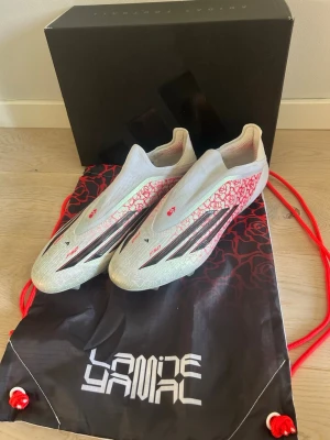 Lamine Yamal F50 - Säljer ett par stilrena fotbollsskor från Adidas i vitt med röda och svarta detaljer. Skorna har en strömlinjeformad design utan snörning och mönstrad ovandel. Perfekta för dig som vill sticka ut på planen. Levereras med matchande svart och röd påse. Säljer dom eftersom att min fot inte passar bra i adidas. Har använt dom 5-10 gånger MAX!