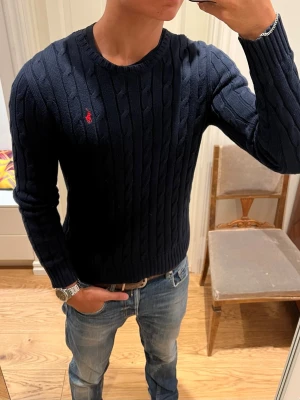 Mörkblå kabelstickad Ralph Lauren Tröja - Kabelstickad mörkblå tröja från Polo Ralph Lauren. Inga defekter. Storlek S.   Axelbredd: 44cm. Övre axelsöm till ärmens slut: 63cm. Övre axelsöm ner till slut: 59cm. Tänk på att tyget är stretchigt och kan passa större. Skriv gärna om du undrar något🙌