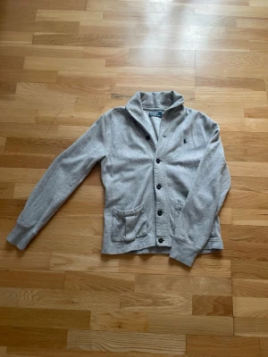 Grå kofta från Polo Ralph Lauren - Snygg grå kofta från Polo Ralph Lauren med knappar framtill, hög krage och två fickor. Klassisk broderad logga på bröstet. Tillverkad i mjukt sweatshirtmaterial som passar perfekt för kyliga dagar.