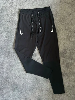 Svarta Nike aeroswift snörning - Säljer ett par svarta mjukisbyxor från Nike med vita loggor på båda sidor framtill. Byxorna har resår i midjan, snörning med Nike-tryck och ribbade muddar vid bensluten. Perfekta för chill eller träning.
