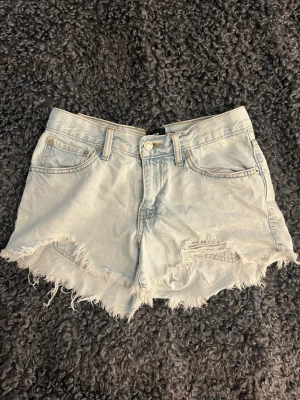 Ljusblå denimshorts med fransig kant - Säljer ett par ljusblå denimshorts med fransig, rå kant och slitna detaljer framtill. Klassisk femficksmodell med knapp och dragkedja. Perfekta för sommaren och ger en avslappnad vibe till din outfit.