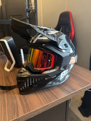 Svart crosshjälm med goggles - Säljer en svart crosshjälm med vita och grå detaljer samt tillhörande goggles med rödtonat spegelglas. Hjälmen har en skärm framtill och ventilationshål. Goggles är från raven.