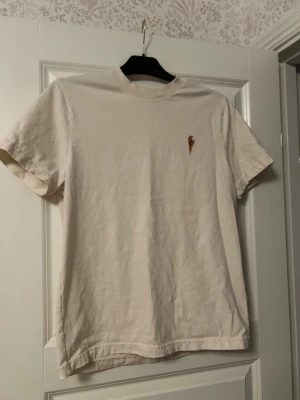 Beige t-shirt med tip top - En stilren beige t-shirt i bomull med korta ärmar och en liten glass på bröstet. Perfekt för dig som gillar enkla plagg med en rolig detalj. Skön passform och passar till det mesta.