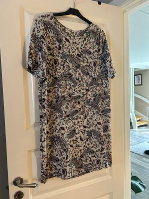 Mönstrad kortklänning med paisley - Kortklänning med paisleyinspirerat mönster i blått, vinrött och svart på vit botten. Klänningen har rund halsringning och korta ärmar. Tillverkad i ett mjukt och lätt material, perfekt för varmare dagar.