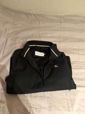 Svart vindjacka från Calvin Klein - Snygg svart vindjacka från Calvin Klein med dragkedja framtill och två fickor med dragkedja. Jackan har vit piping vid kragen och diskret CK-logga på bröstet. Tillverkad i lätt och smidigt material, perfekt för blåsiga dagar.