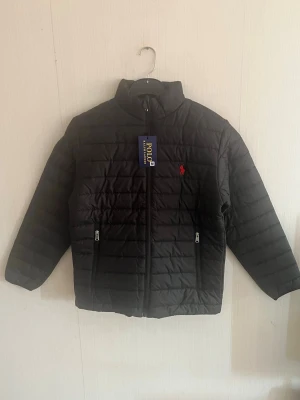 Svart dunjacka från Polo Ralph Lauren - Snygg svart dunjacka med diskret quiltad design och röd logga på bröstet. Jackan har hög krage, två dragkedjeförsedda fickor och smidig dragkedja framtill. Perfekt för skolan och höstdagarna och riktigt stilren look.