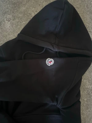 Svart Moncler hoodie med logga - Säljer en svart hoodie från Moncler med stor vit och röd logga på bröstet och en mindre patch på ärmen. Hoodien har huva med dragsko och en klassisk känguruficka framtill. Tillverkad i mjukt material som känns skönt mot huden.