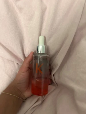 Kérastase hårserum med pipett - Säljer ett Kérastase hårserum i genomskinlig glasflaska med silvrigt lock och vit pipett. Innehållet är orangefärgat och flaskan har en stilren design med stor orange K på framsidan. Perfekt för dig som vill boosta håret med extra vård.