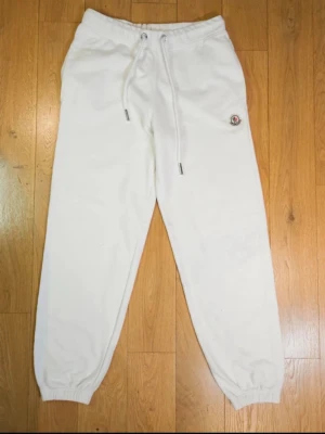 Vita mjukisbyxor från Moncler - Säljer ett par vita mjukisbyxor från Moncler med dragsko i midjan och ribbade muddar vid bensluten. Byxorna har en liten Moncler-logga på ena benet och är tillverkade i mjukt material som känns riktigt skönt mot huden.