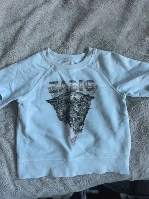 Ljusblå sweatshirt Zadig & Voltaire - Ljusblå sweatshirt från Zadig & Voltaire med ett stort glittrigt pantermotiv och texten 'ZADIG' framtill. Tröjan har rund halsringning och långa ärmar. Materialet är mjukt och bekvämt, perfekt för en avslappnad stil.