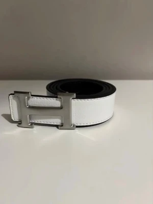 Vitt skärp med silverfärgat H-spänne - Snyggt vitt bälte med ett stort H-format spänne i silverfärg. Bältet har en stilren och modern look, perfekt för att lyfta vilken outfit som helst. Tillverkat i skinn med slät yta och svarta kanter för extra kontrast.