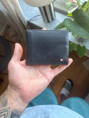 Svart läderplånbok Tommy Hilfiger - Snygg svart plånbok i äkta läder från Tommy Hilfiger. Klassisk design med flera kortfack och ett fack för sedlar. Plånboken har en liten metallogo på framsidan och en randig detalj på insidan. Perfekt storlek för fickan.