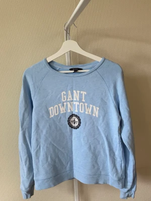 Ljusblå sweatshirt från GANT - Ljusblå sweatshirt från GANT med vit text 'GANT DOWNTOWN' och ett tryckt emblem på bröstet. Tröjan har rund halsringning, långa ärmar och ribbade muddar. Perfekt för en avslappnad och sportig stil. Se fläck men det syns inte så mycket. 