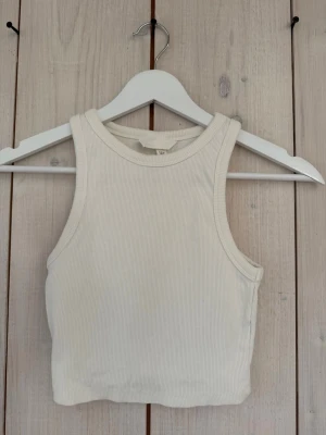 Vit ribbad linne/topp från H&M - Säljer en fräsch vit ribbad topp från H&M i storlek XS. Toppen är ärmlös och croppad med en klassisk rund halsringning. Perfekt att styla till jeans eller kjol för en clean och enkel look.