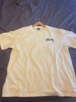 Beige Stüssy t-shirt med tärningsmotiv - Vit t-shirt från Stüssy med svart logga på bröstet och stort tryck på ryggen med lila tärningar och Stüssy-logga. Klassisk passform och rund hals. Perfekt för dig som gillar streetwear och vill sticka ut lite extra.