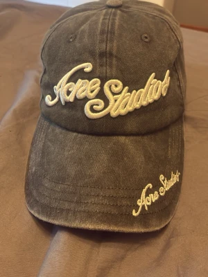 Svart keps från Acne Studios - Säljer en svart keps från Acne Studios med broderad logga i vit text både framtill och på skärmen. Kepsen är tillverkad i 100% bomull och har en klassisk, böjd skärm och justerbar passform. Perfekt för en trendig och avslappnad stil.
