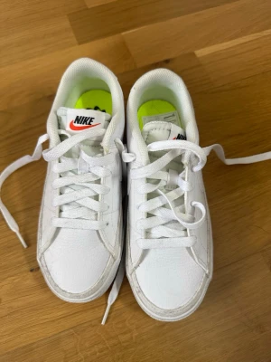 Vita Nike sneakers med låg profil - Fräscha vita Nike sneakers med klassisk låg siluett och vit sula med svart rand. Skorna har vita skosnören, diskret swoosh på sidan och en neonfärgad innersula. Tillverkade i läder med textildetaljer och passar perfekt till streetwear.