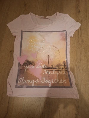 Ljusrosa t-shirt med hjärtan och tryck - Säljer en ljusrosa t-shirt med ett stort tryck framtill som visar ett pariserhjul, rosa hjärtan och texten 'Thinking about your smile, the lights, Always Together'. T-shirten har rund hals och korta ärmar. Perfekt för dig som gillar gulliga motiv och mjuka färger.