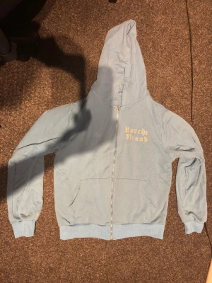 Ljusblå Rinestone hoodie - Säljer en ljusblå hoodie med dragkedja och huva. På bröstet står det 'Sorche Brand' i gul text och på ryggen finns en stor ros i glittriga stenar. Hoodien har fickor framtill och ribbade muddar vid ärmar och midja. Perfekt för dig som gillar unika detaljer!