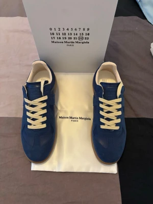 Blå Maison Margiela sneakers - Snygga blå sneakers från Maison Martin Margiela med vita snören och beige innersula. Skorna har en klassisk låg siluett och ovandel i mocka och läder. Yttersulan är i gummi för bra grepp. Perfekta för dig som vill ha något stilrent och unikt.
