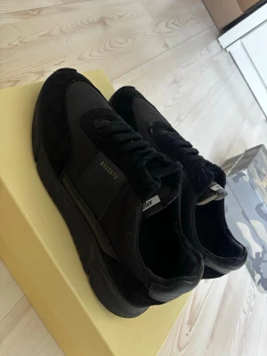 Axel Arigato skor - Snygga helsvarta Axel Arigato sneakers i stilren och exklusiv design. Skorna är använda och har en liten solblekning/solbränna, vilket syns lite på färgen. I övrigt är de fortfarande riktigt snygga och passar perfekt till både jeans och streetwear.
